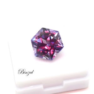 Puede incluir: Una piedra preciosa hexagonal facetada que muestra un cambio de color vibrante entre tonos p&uacute;rpura intenso y magenta. La piedra est&aacute; colocada sobre una base blanca con el texto negro Binzal impreso en la esquina inferior izquierda.