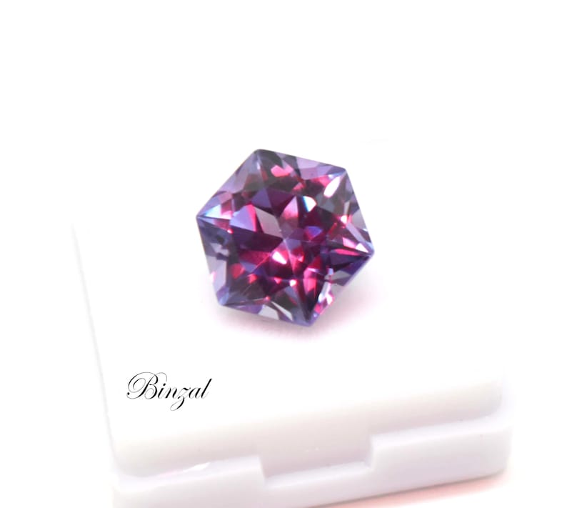 Puede incluir: Una piedra preciosa hexagonal facetada que muestra un cambio de color vibrante entre tonos p&uacute;rpura intenso y magenta. La piedra est&aacute; colocada sobre una base blanca con el texto negro Binzal impreso en la esquina inferior izquierda.