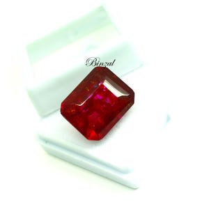 May include: A striking, rectangular-cut ruby gemstone, displaying a rich red colour. The gem is presented against a white backdrop, highlighting its facets and sparkle. The word "Binzal" is written in cursive on the top left.