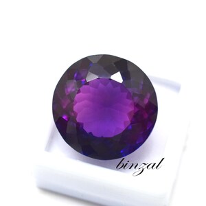 May include: A large, round, faceted amethyst gemstone with a deep purple hue. The gem is set against a white background, showcasing its brilliant cut and color. The word "binzal" is printed on the white surface.