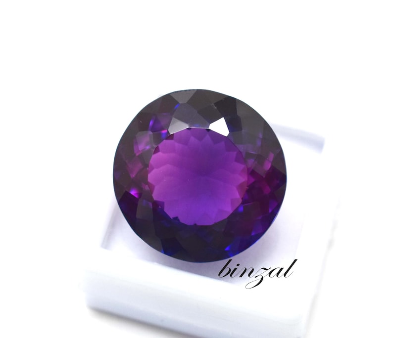 May include: A large, round, faceted amethyst gemstone with a deep purple hue. The gem is set against a white background, showcasing its brilliant cut and color. The word "binzal" is printed on the white surface.