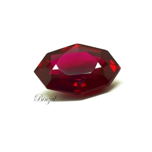 May include: A faceted, deep red gemstone with a unique octagonal shape. The gem has multiple facets that reflect light, creating a brilliant sparkle. The word "Binzal" is written in cursive script on the lower left.