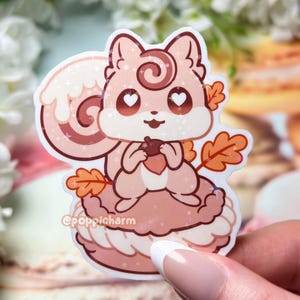 May include: A sticker featuring a cartoon squirrel holding an acorn, sitting on a pink dessert. The squirrel is light brown with heart-shaped eyes and autumn leaves. The sticker has the text "@poppicharm".