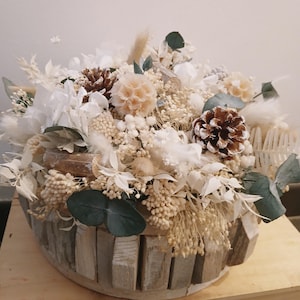 Pode incluir: Arranjo floral decorativo com flores secas, pinhas e folhas de eucalipto. O arranjo está numa cesta de madeira rústica. A paleta de cores inclui tons de branco, bege e castanho, criando uma estética natural e elegante.
