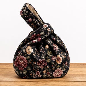 Può includere: Una borsa in tessuto nero con chiusura a nodo e motivo floreale nei toni del rosso scuro, rosa e verde tenue. La borsa ha una forma arrotondata e arricciata con un manico unico formato passando un lato nell'altro e una fodera interna chiara.