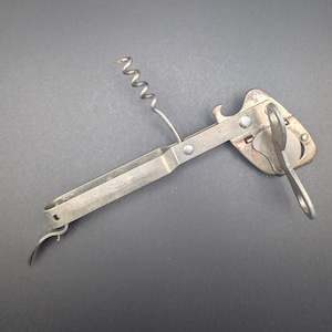 Pode incluir: Um utensílio de cozinha vintage multi-ferramentas. A ferramenta prateada combina um saca-rolhas, um abridor de garrafas e um abridor de latas. O saca-rolhas tem um design em espiral. O abridor de latas tem um design em forma de chave. A ferramenta está numa superfície cinza escuro.