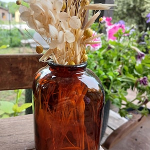 Puede incluir: Un jarrón de cristal marrón lleno de flores y hierbas secas. El jarrón tiene una forma ancha y redondeada y un color ámbar oscuro. Las flores secas son de color blanco crema y las hierbas son de color beige claro. El arreglo está sobre una superficie de madera.