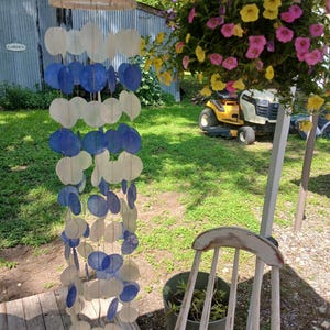 Könnte beinhalten: Ein hängendes Windspiel mit abwechselnd weißen und blauen kreisförmigen Elementen. Das Windspiel ist an einem kreisförmigen Holzrahmen aufgehängt. Ein hängender Korb mit rosa und gelben Blumen ist im Hintergrund zu sehen, zusammen mit einem weißen Stuhl.