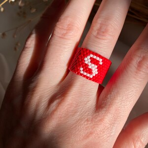 Op de afbeelding: Een rode kralenring met een wit "S"-ontwerp. De ring is gemaakt van kleine, gelijkmatig verdeelde kralen. Een opvallend en opvallend accessoire.