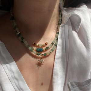 Puede incluir: Un collar de varias hebras con cuentas verdes y doradas, y un colgante de estrella dorada. El collar tiene un diseño delicado con una mezcla de tonos verdes y dorados. El collar se lleva sobre una camisa blanca.