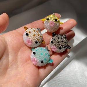 May include: Four spherical pufferfish charms crafted from small beads. Each charm features a round body, small fins, black bead eyes, and a pink beaded mouth. The charms are colored in yellow, white with brown spots, grey with black spots, and light blue.