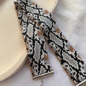May include: A beaded bracelet with a snake skin pattern in black, white, gray, and tan. The bracelet has a silver clasp and is laying on a white surface.