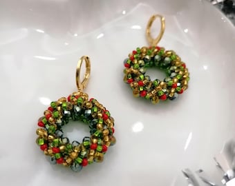 May include: Gold-coloured hoop earrings, each with a beaded wreath design. The wreaths are made of green, red, and gold beads, giving a festive holiday appearance. The earrings are presented on a white surface.