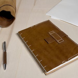 Può includere: Un diario in pelle marrone con chiusura a cinturino e cuciture bianche. Una penna abbinata con dettagli argentati è accanto al diario. Sono presenti anche un rotolo di pelle e una pila di carta bianca.
