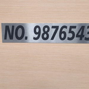 May include: A rectangular, brushed metal sign with the text "NO. 9876543" in black, mounted on a light wood-grain surface. The sign is a simple, modern design, suitable for a house number or office signage.