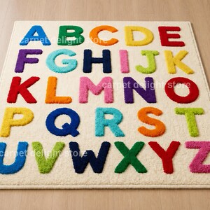 May include: A rectangular area rug with a cream-colored background featuring the English alphabet in raised, multi-colored letters. Each letter is rendered in a distinct, vibrant hue including blue, green, orange, yellow, red, purple, and pink.