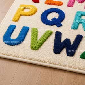 May include: A cream-colored area rug featuring a tufted alphabet design with raised, textured letters in various colors including yellow, blue, green, orange, pink, and dark blue. The letters U, V, W, X, Q, and R are clearly visible on the surface.