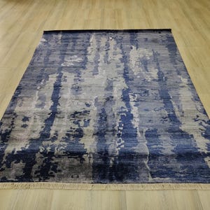 Puede incluir: Una alfombra rectangular con un patr&oacute;n abstracto y moteado en tonos azul marino, azul claro y gris. La alfombra tiene una superficie texturizada y est&aacute; rematada con flecos cortos de color claro en los bordes superior e inferior.