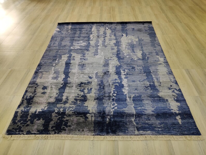 Puede incluir: Una alfombra rectangular con un patr&oacute;n abstracto y moteado en tonos azul marino, azul claro y gris. La alfombra tiene una superficie texturizada y est&aacute; rematada con flecos cortos de color claro en los bordes superior e inferior.