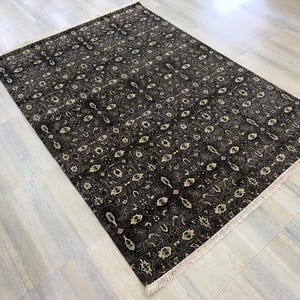 Puede incluir: Una alfombra rectangular con un fondo gris oscuro y negro que presenta un patr&oacute;n geom&eacute;trico y floral repetitivo en color crema. La alfombra tiene una textura de pelo corto e incluye flecos de color claro en los dos bordes m&aacute;s cortos.