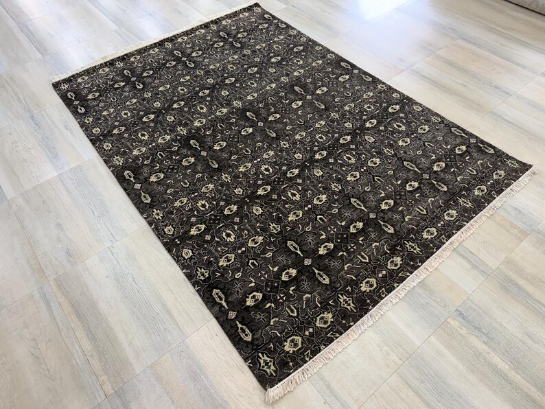 Puede incluir: Una alfombra rectangular con un fondo gris oscuro y negro que presenta un patr&oacute;n geom&eacute;trico y floral repetitivo en color crema. La alfombra tiene una textura de pelo corto e incluye flecos de color claro en los dos bordes m&aacute;s cortos.