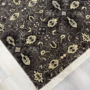 Puede incluir: Una alfombra rectangular con un fondo gris carb&oacute;n oscuro y un patr&oacute;n geom&eacute;trico repetitivo en crema y beige claro. La alfombra tiene un pelo denso y texturizado, y est&aacute; rematada con una hilera de flecos blancos cortos en el borde.