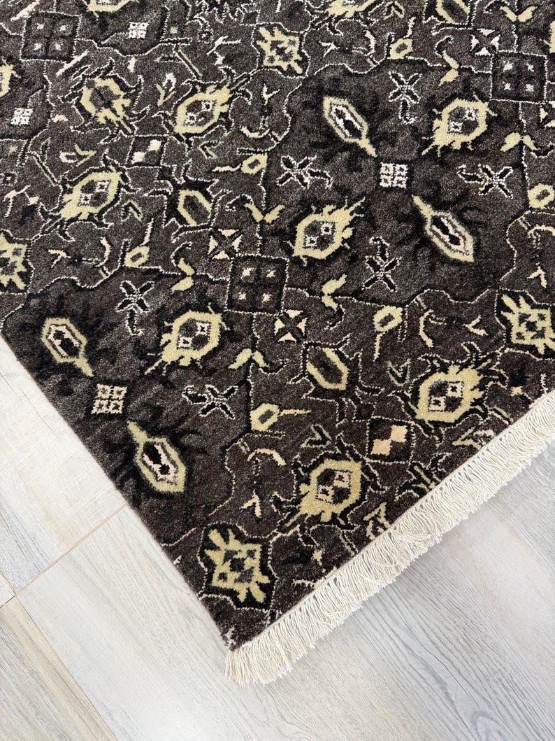 Puede incluir: Una alfombra rectangular con un fondo gris carb&oacute;n oscuro y un patr&oacute;n geom&eacute;trico repetitivo en crema y beige claro. La alfombra tiene un pelo denso y texturizado, y est&aacute; rematada con una hilera de flecos blancos cortos en el borde.