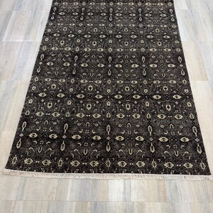 Puede incluir: Una alfombra rectangular con fondo gris oscuro y un patr&oacute;n geom&eacute;trico repetitivo en crema y gris claro. La alfombra presenta un dise&ntilde;o denso y sim&eacute;trico de motivos florales y de diamantes estilizados, rematada con flecos blancos.