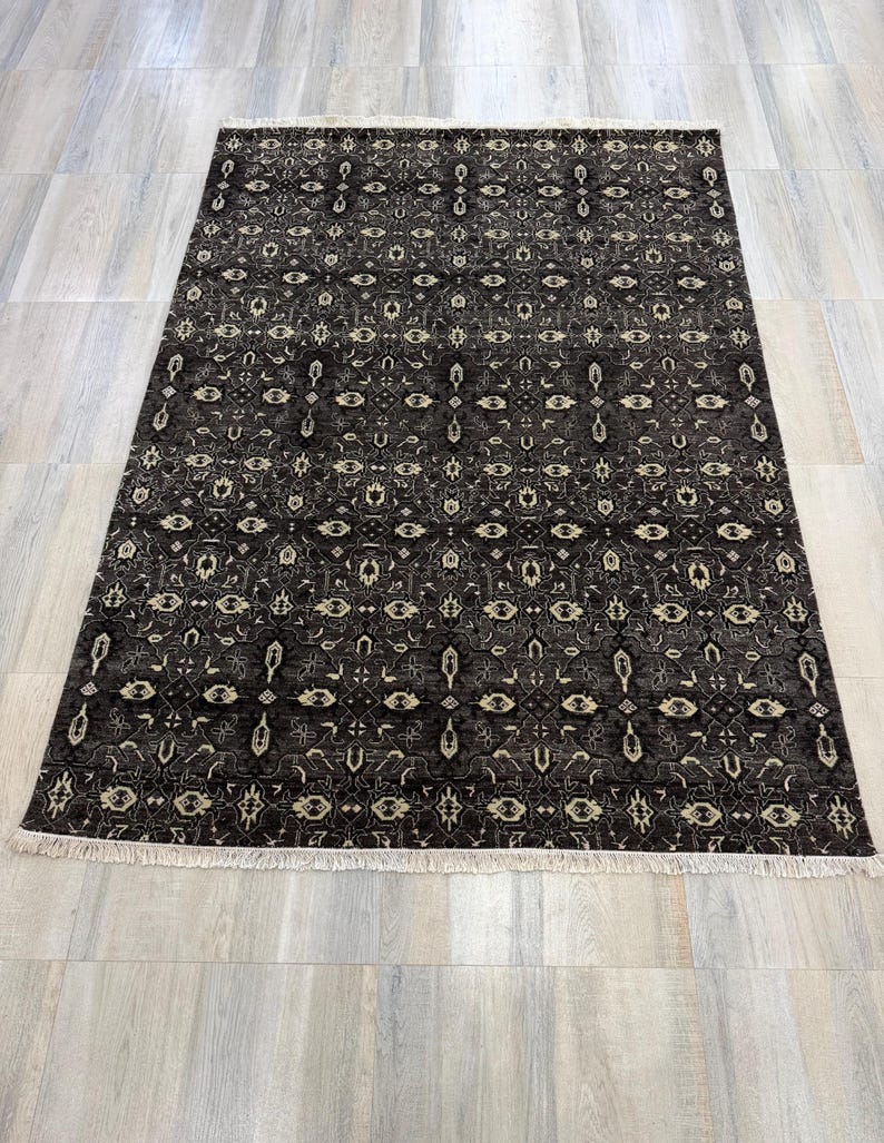 Puede incluir: Una alfombra rectangular con fondo gris oscuro y un patr&oacute;n geom&eacute;trico repetitivo en crema y gris claro. La alfombra presenta un dise&ntilde;o denso y sim&eacute;trico de motivos florales y de diamantes estilizados, rematada con flecos blancos.