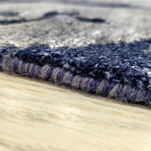 Puede incluir: Una vista de cerca del borde de una alfombra que muestra un pelo grueso y texturizado en tonos azul marino, gris y blanco. El borde est&aacute; rematado con una puntada de sobrehilado densa con hilo azul oscuro y gris, resaltando la construcci&oacute;n.