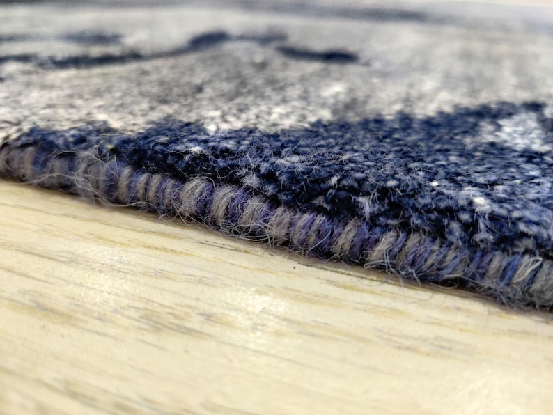 Puede incluir: Una vista de cerca del borde de una alfombra que muestra un pelo grueso y texturizado en tonos azul marino, gris y blanco. El borde est&aacute; rematado con una puntada de sobrehilado densa con hilo azul oscuro y gris, resaltando la construcci&oacute;n.