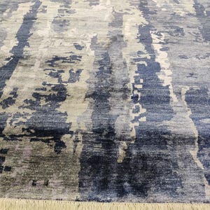 Puede incluir: Una alfombra rectangular con un patr&oacute;n abstracto de rayas verticales en tonos azul marino, azul pizarra, gris claro y blanco. El dise&ntilde;o incluye parches texturizados de aspecto desgastado y un fleco en el borde inferior.