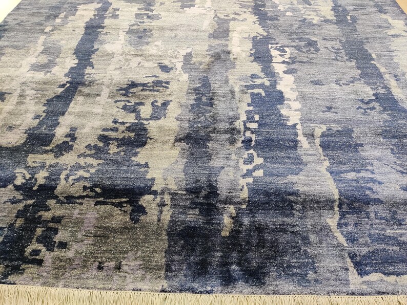 Puede incluir: Una alfombra rectangular con un patr&oacute;n abstracto de rayas verticales en tonos azul marino, azul pizarra, gris claro y blanco. El dise&ntilde;o incluye parches texturizados de aspecto desgastado y un fleco en el borde inferior.