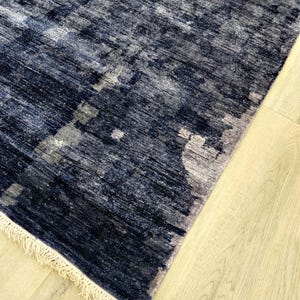 Puede incluir: Una alfombra rectangular con un patr&oacute;n abstracto en azul marino profundo y gris carb&oacute;n con una apariencia texturizada y desgastada. La alfombra est&aacute; terminada con flecos cortos de color crema en el borde inferior.