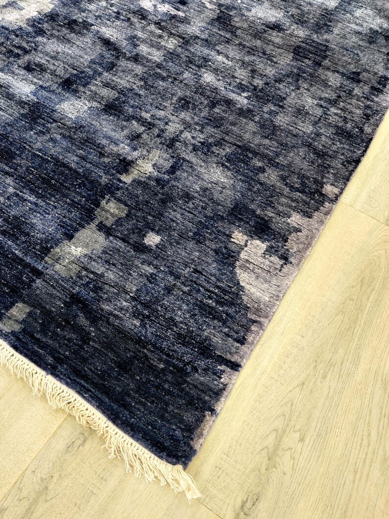 Puede incluir: Una alfombra rectangular con un patr&oacute;n abstracto en azul marino profundo y gris carb&oacute;n con una apariencia texturizada y desgastada. La alfombra est&aacute; terminada con flecos cortos de color crema en el borde inferior.