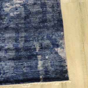 Puede incluir: Una alfombra rectangular con una paleta de colores azul marino profundo e &iacute;ndigo con parches abstractos y moteados de gris claro y blanco. La alfombra tiene una superficie tejida texturizada y un fleco corto de color blanco roto en el borde inferior.