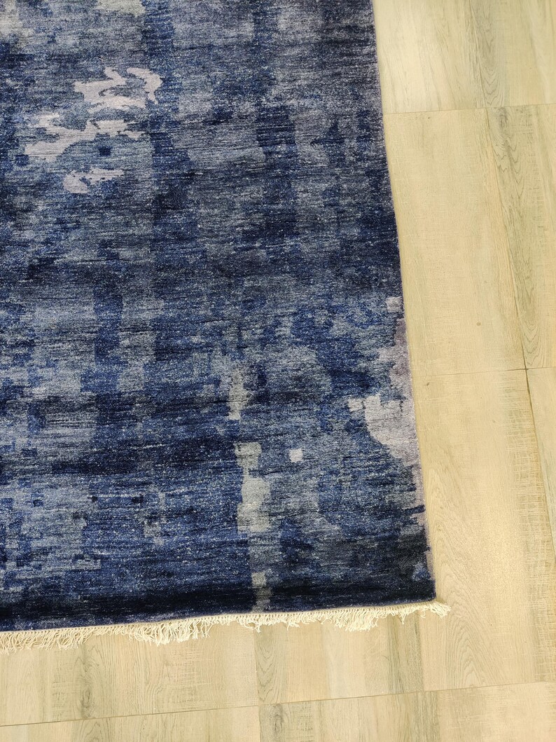 Puede incluir: Una alfombra rectangular con una paleta de colores azul marino profundo e &iacute;ndigo con parches abstractos y moteados de gris claro y blanco. La alfombra tiene una superficie tejida texturizada y un fleco corto de color blanco roto en el borde inferior.