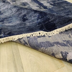 Puede incluir: Una alfombra rectangular con un patr&oacute;n abstracto en tonos azul marino profundo, gris pizarra y blanco. La alfombra tiene una esquina doblada que revela una superficie texturizada y est&aacute; rematada con flecos de color blanco roto.