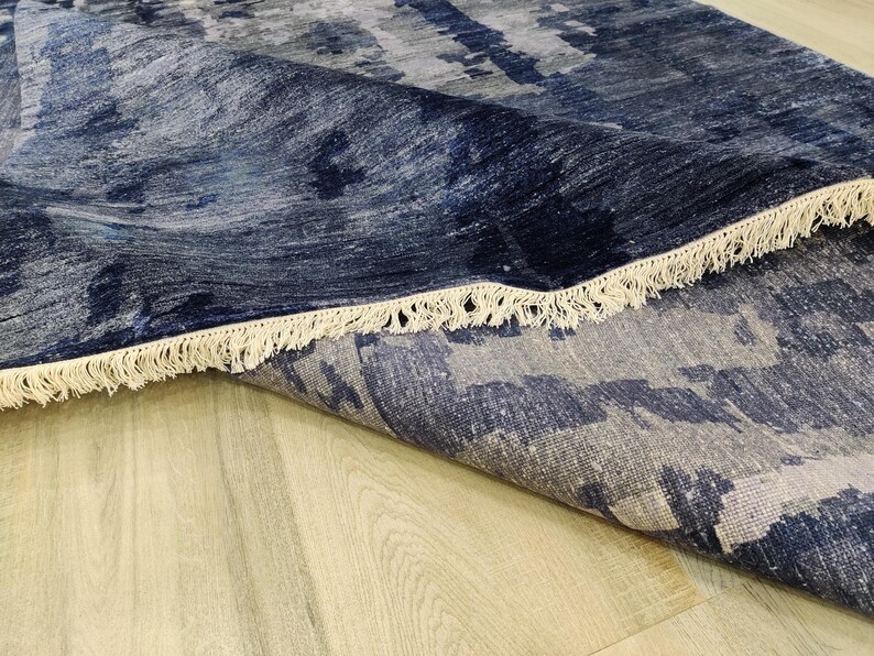 Puede incluir: Una alfombra rectangular con un patr&oacute;n abstracto en tonos azul marino profundo, gris pizarra y blanco. La alfombra tiene una esquina doblada que revela una superficie texturizada y est&aacute; rematada con flecos de color blanco roto.