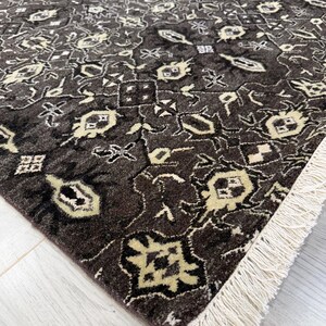Puede incluir: Una alfombra rectangular con fondo marr&oacute;n oscuro y un patr&oacute;n geom&eacute;trico repetitivo en crema y beige claro. La alfombra tiene un pelo denso y texturizado, y est&aacute; rematada con una hilera de flecos de color blanco roto en el borde corto.