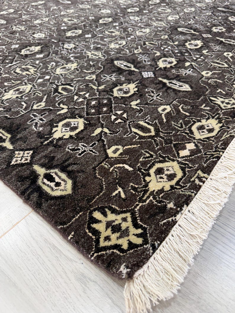 Puede incluir: Una alfombra rectangular con fondo marr&oacute;n oscuro y un patr&oacute;n geom&eacute;trico repetitivo en crema y beige claro. La alfombra tiene un pelo denso y texturizado, y est&aacute; rematada con una hilera de flecos de color blanco roto en el borde corto.