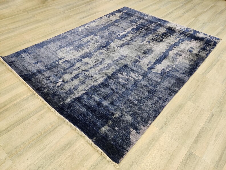 Puede incluir: Una alfombra rectangular con un patr&oacute;n abstracto en tonos azul marino, gris pizarra y blanco. El dise&ntilde;o consiste en bandas horizontales texturizadas superpuestas y parches moteados. La alfombra tiene flecos cortos de color claro en los extremos.