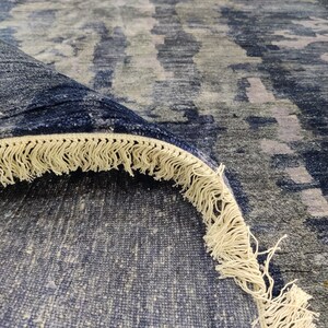 Puede incluir: Una alfombra grande con un patr&oacute;n abstracto en tonos azul marino profundo, gris pizarra y gris claro. La alfombra est&aacute; doblada para mostrar su parte inferior y un grueso fleco de color crema a lo largo del borde de la tela tejida.