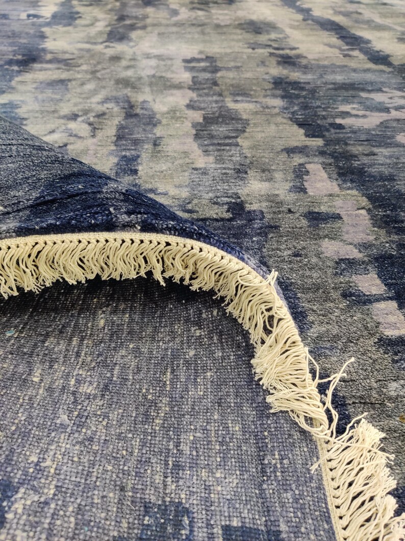 Puede incluir: Una alfombra grande con un patr&oacute;n abstracto en tonos azul marino profundo, gris pizarra y gris claro. La alfombra est&aacute; doblada para mostrar su parte inferior y un grueso fleco de color crema a lo largo del borde de la tela tejida.