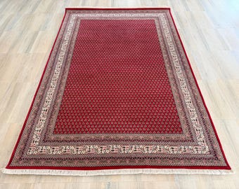 Puede incluir: Una alfombra rectangular con un campo central de color rojo intenso con un patrón repetitivo a pequeña escala. La alfombra está enmarcada por varios bordes decorativos en tonos crema, negro y rojo, con flecos blancos cortos en los extremos.