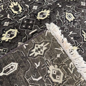 Puede incluir: Una alfombra de color gris carb&oacute;n oscuro con un patr&oacute;n geom&eacute;trico repetitivo en blanco crema. La alfombra tiene un pelo denso y texturizado, y un borde acabado con largos flecos de color crema que se extienden desde un lado del tejido.