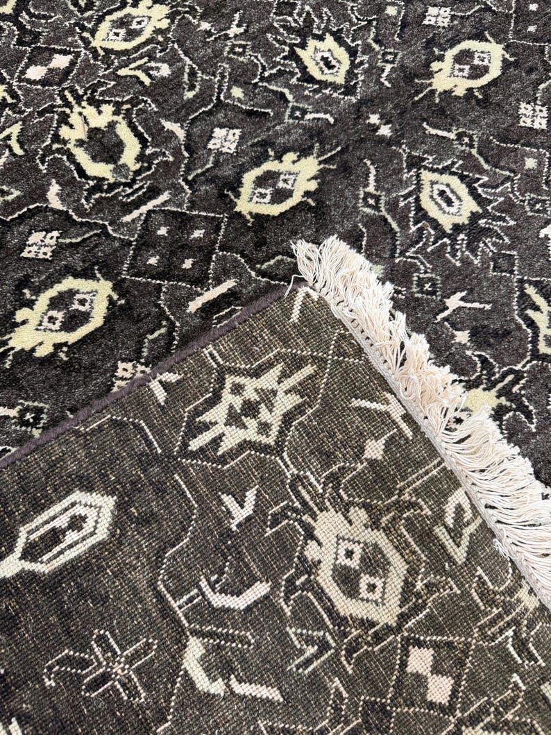 Puede incluir: Una alfombra de color gris carb&oacute;n oscuro con un patr&oacute;n geom&eacute;trico repetitivo en blanco crema. La alfombra tiene un pelo denso y texturizado, y un borde acabado con largos flecos de color crema que se extienden desde un lado del tejido.