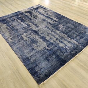 Puede incluir: Una alfombra rectangular con un patr&oacute;n abstracto desgastado en tonos azul marino profundo, gris pizarra y blanco. La alfombra tiene una superficie texturizada con flecos visibles en los bordes cortos, mostrando un efecto de color en capas.