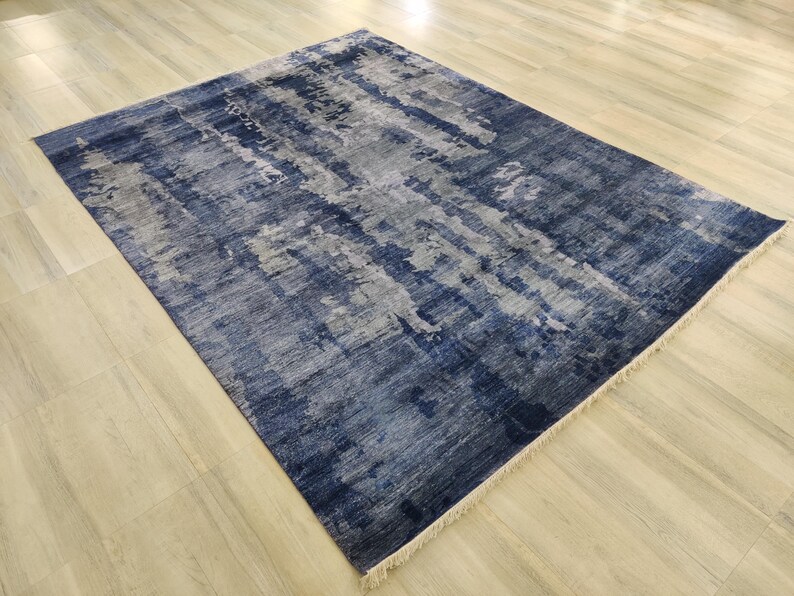 Puede incluir: Una alfombra rectangular con un patr&oacute;n abstracto desgastado en tonos azul marino profundo, gris pizarra y blanco. La alfombra tiene una superficie texturizada con flecos visibles en los bordes cortos, mostrando un efecto de color en capas.