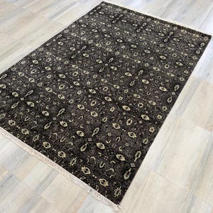 Puede incluir: Una alfombra rectangular con un fondo gris carb&oacute;n oscuro y un patr&oacute;n repetitivo de motivos florales geom&eacute;tricos en color crema y beige claro. La alfombra tiene una textura de pelo corto y est&aacute; rematada con flecos claros en ambos extremos.